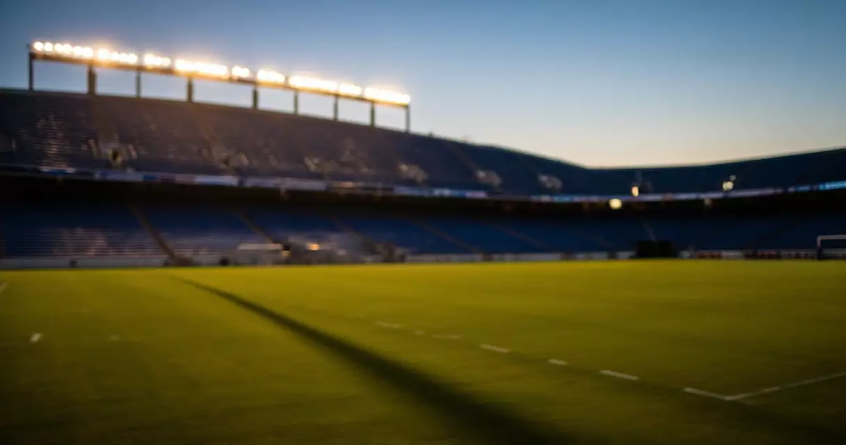 Empty football stadium at dusk with lights on and natural shadows across the field