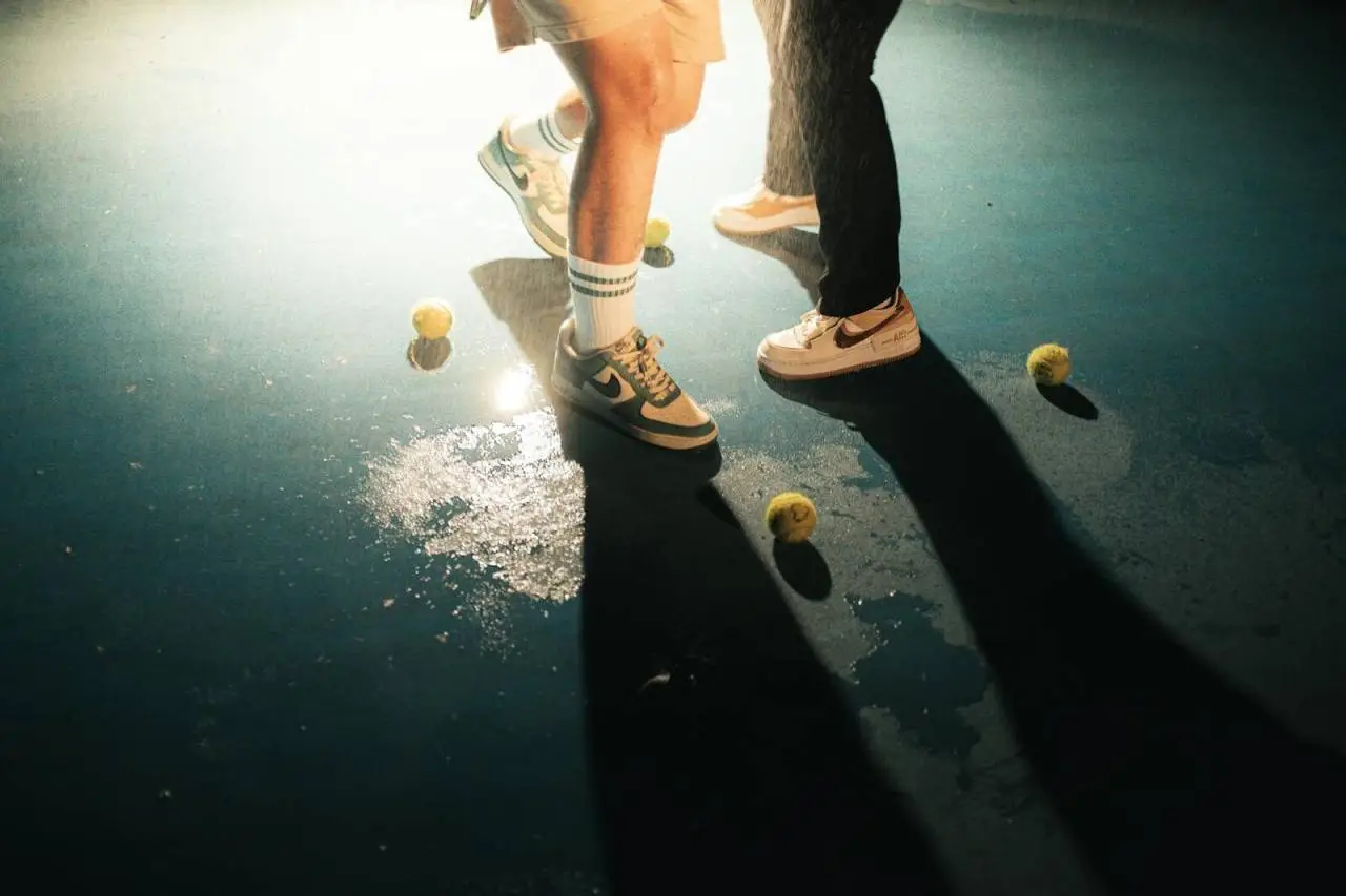 Close-up of tennis shoes and balls on wet court with dramatic lighting.